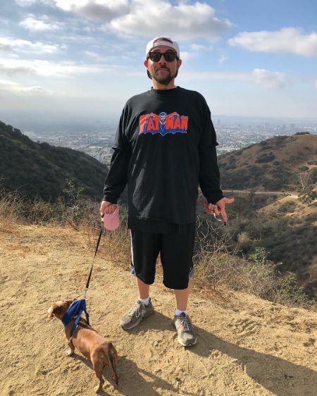 Kevin Smith in a black t-shirt and shorts poses at a hill with his dog.